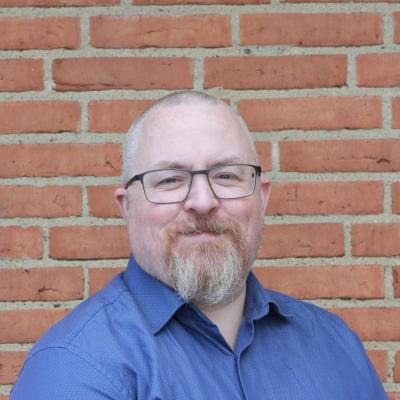 Close-up photo of old white man in a blue shirt and with greying beard, very little hair and glasses standing in front of a red brick wall.
